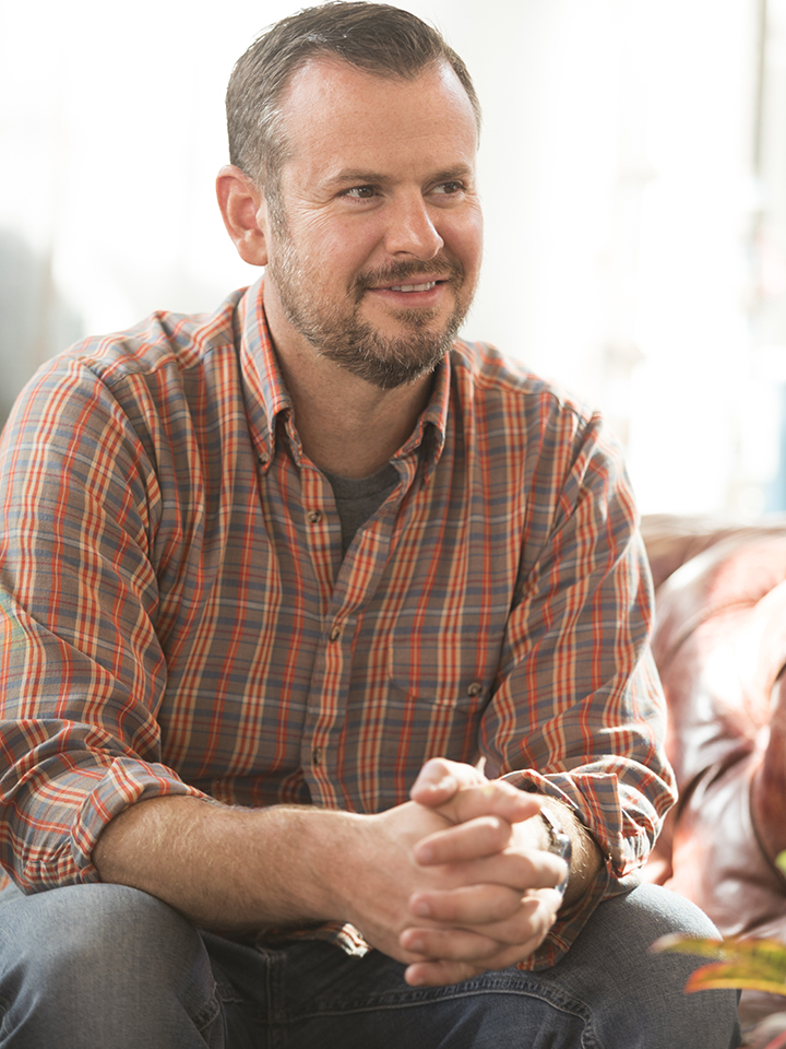 photo of a smiling bearded man with short brown hair, wearing an orange plaid shirt looking off camera.