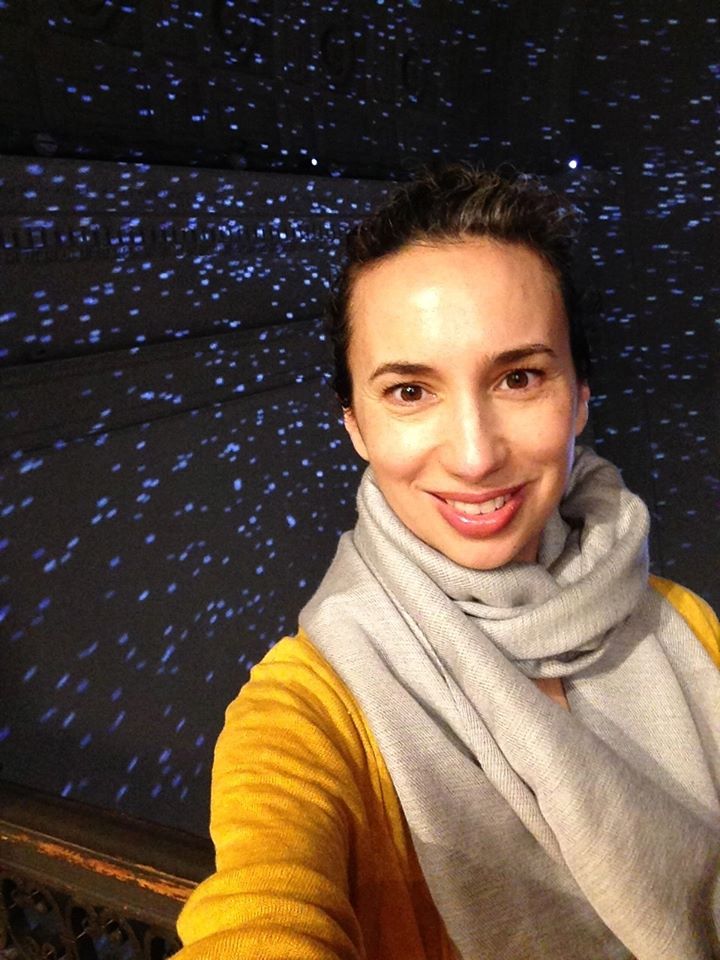 Photo of a smiling woman with dark curly hair wearing a yellow sweater and a beige scarf against a dark starry background.