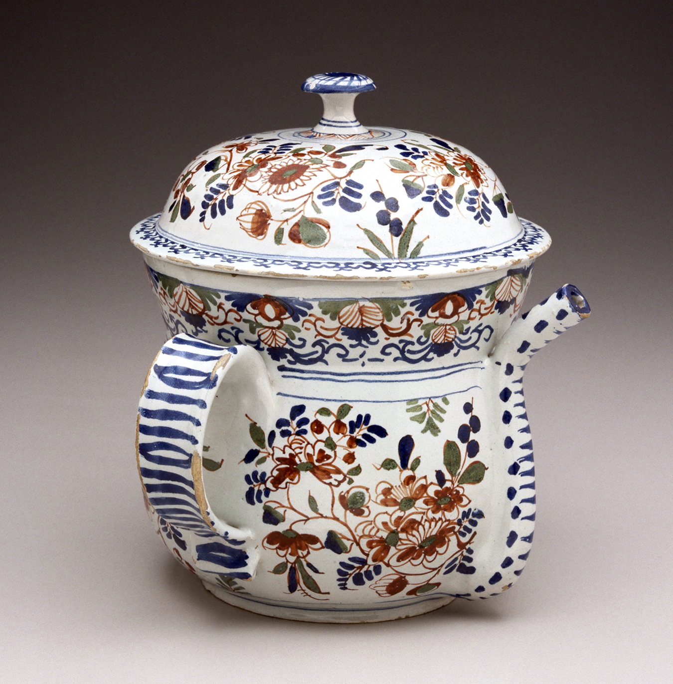 A small white lidded pot with a spout and handle. The white surface is painted with red, green, and blue decorations.